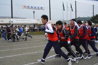 滑川町総合運動公園でおこなわれた町民体育祭でお互いの両足を白色の紐で結びオレンジ色のゼッケンを着て真剣な表情でムカデ競争をする5人の男性の写真